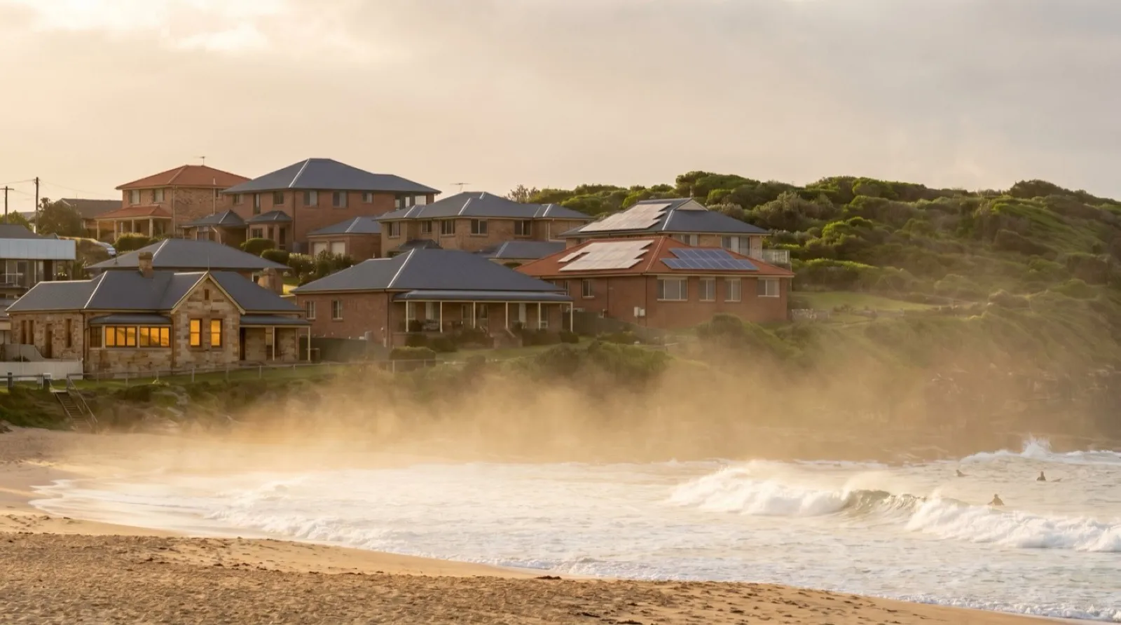 Golden-hour view along a Newcastle surf beach, salt spray hazing up off the breaking waves toward established brick-and-Colorbond houses on the low headland behind, a couple of them with rooftop solar panels